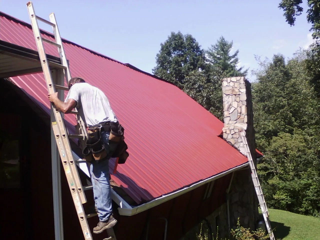 Roofer on a ladder beside a red metal roof and stone chimney — Soffit & Fascia Repair by Roofing Brothers in Coppell